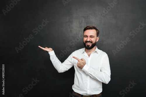 Optimistic bearded man pointing index finger while holding thing on palm, demonstrating or advertising over dark gray background copy space