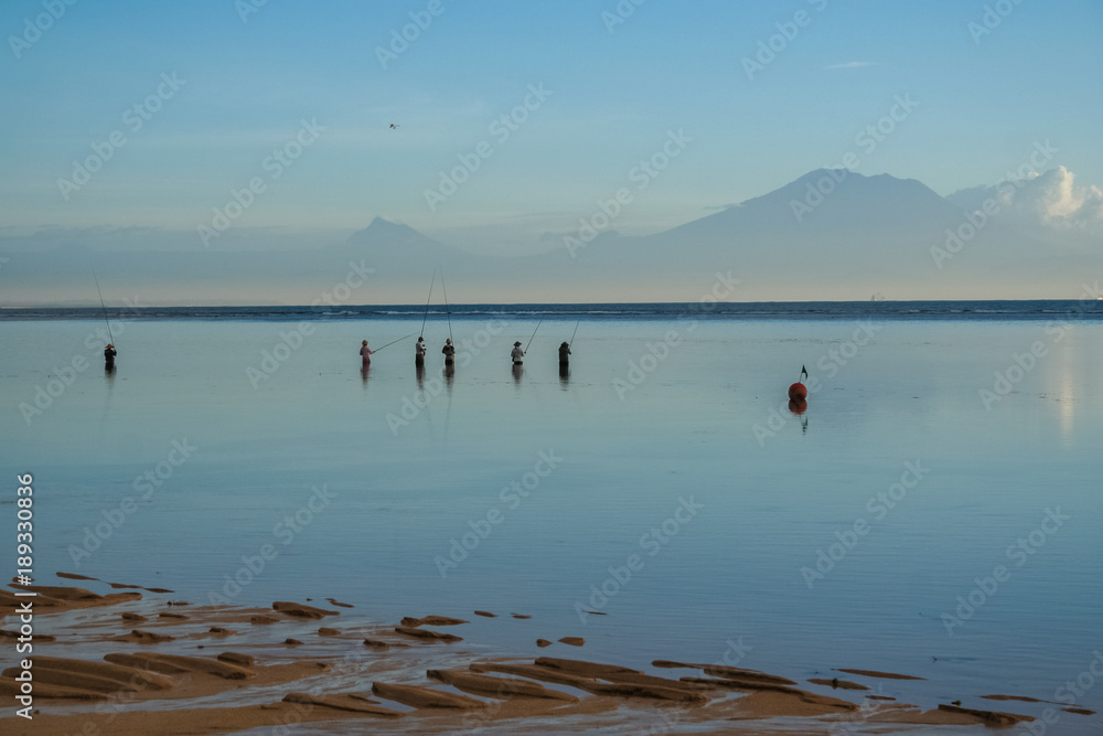 A peaceful image of local fishermen standing in the sea at dawn during low tide, holding their fishing rods and waiting for a catch. Taken at a beach in Bali, Indonesia.  