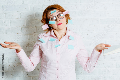 Red hair comic woman in glasses with stickers on her, looking at camera with a quizzical lose courage emotion, and writting in notebook a lot of things to do. White brick wall with shadow behind her. photo