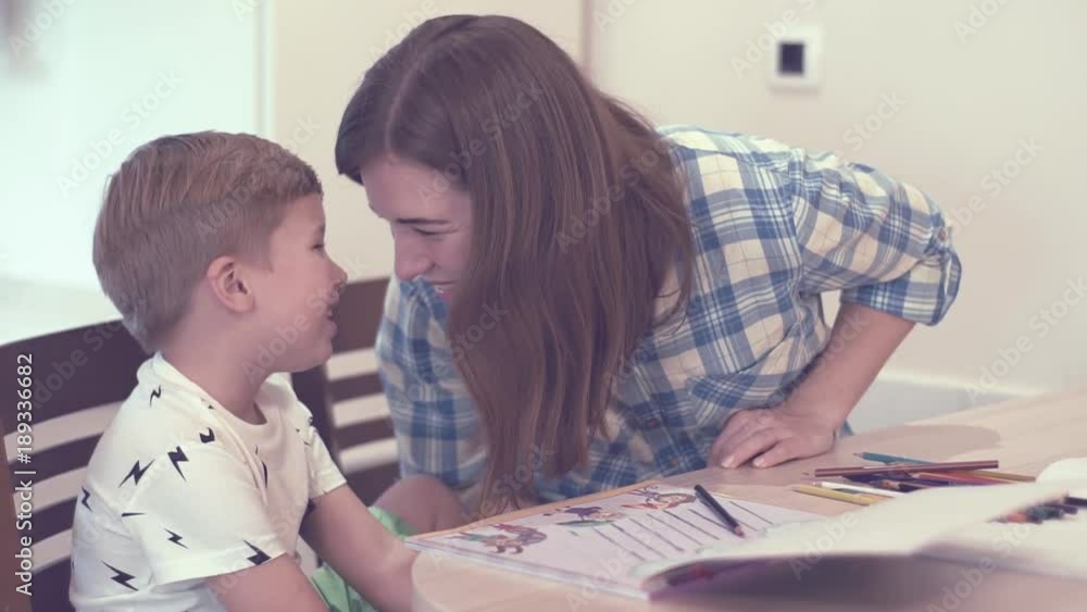 Beautiful Young Mother In Plaid Blue Shirt Smiles And Says Something To Her Little Curious Blurred Son While Doing Homework Sitting At The Table Beautiful Young Mother In Plaid Blue Shirt Smiles And Says Something To Her Little Curious Blurred Son While Doing Homework Sitting At The Table