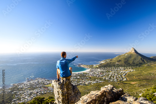 Young male hiker sitting on a rock at Kasteelspoort Hiking Trail in Table Mountain National Park, Cape Town, South Africa, pointing at the suburb of Camps Bay and Lion`s Head mountain in background. photo