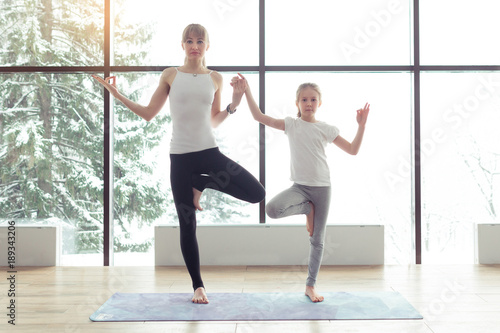 Mother and kid daughter in the gym centre doing yoga poses or stretching fitness exercise near big window with a beautiful scenery with mountains and trees with snow. Healthy family lifestyle concept
