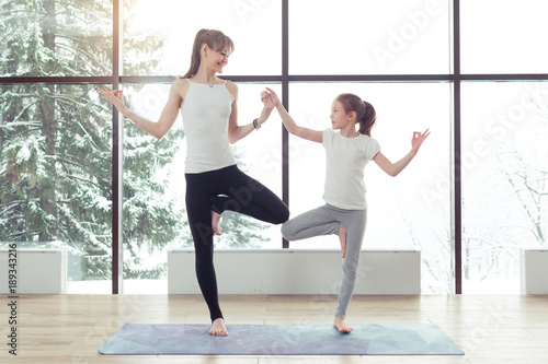 Young beautiful woman and charming little daughter are doing difficult yoga exercises and smiling while working out at gym with beautiful scenery outside window with sunlight. Healthy fitness concept