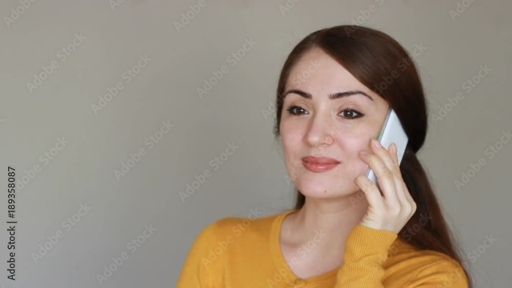 Beautiful young brunette woman talks on the mobile phone, smiling and says i love you. Happy girl with a white smartphone in her hand on a light background looks at the camera. Beautiful young brunette woman talks on the mobile phone, smiling and says i love you. Happy girl with a white smartphone in her hand on a light background looks at the camera.
