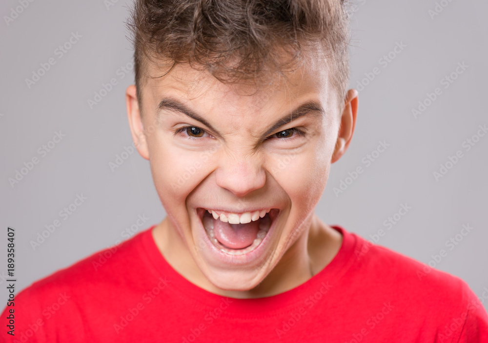 Emotional portrait of irritated shouting teen boy. Furious teenager screaming and looking with anger at camera. Handsome outraged child shouting out loud on gray background. Emotional portrait of irritated shouting teen boy. Furious teenager screaming and looking with anger at camera. Handsome outraged child shouting out loud on gray background.