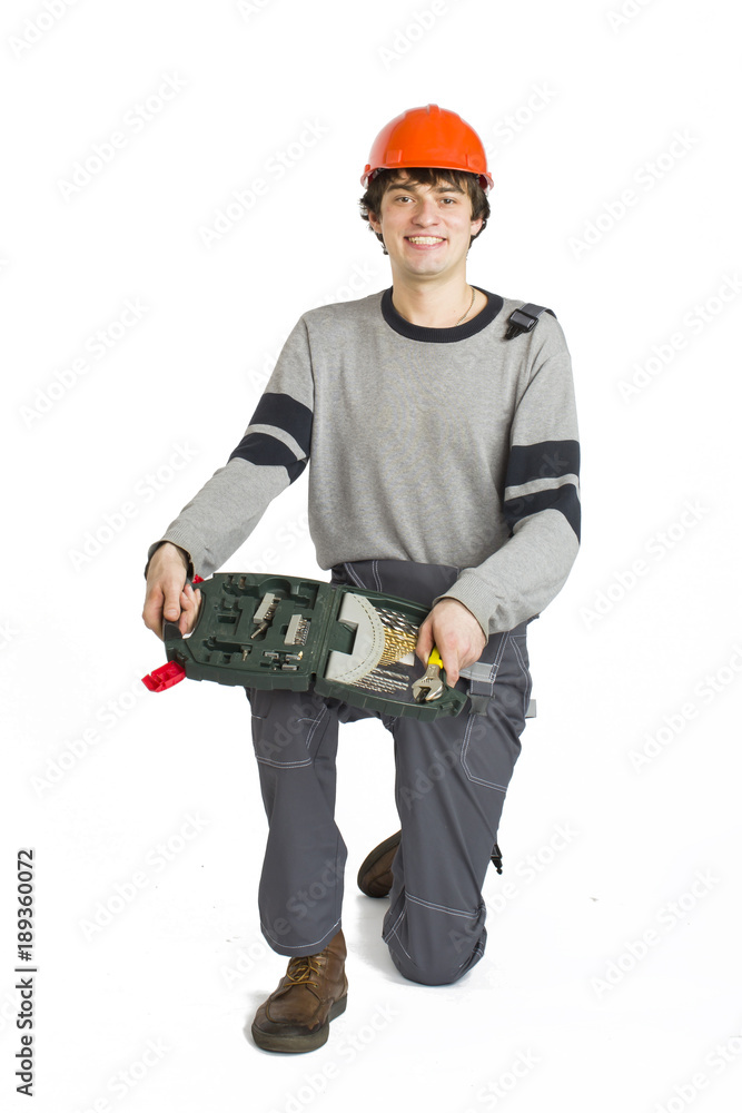 A young man in working grey clothes and orange hard helmet sitting with toolkit on isolated white background. A young man in working grey clothes and orange hard helmet sitting with toolkit on isolated white background.