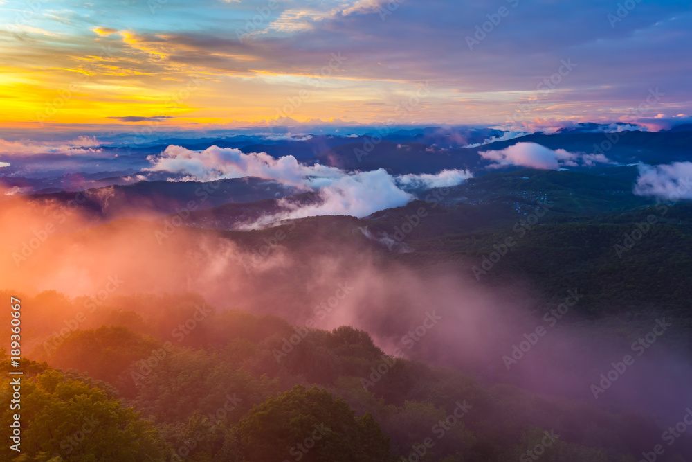 Sunset hidden behind clouds and fog over hills, bright yellow sun on colorful cloudscape, blue violet orange sky. Panorama of the Black Sea coastline from Akhun mountain, Big Sochi, Russia.