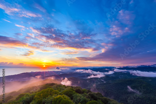 Sunset hidden behind clouds and fog over hills, bright yellow sun on colorful cloudscape, blue violet orange sky. Panorama of the Black Sea coastline from Akhun mountain, Big Sochi, Russia. © Valery Bocman
