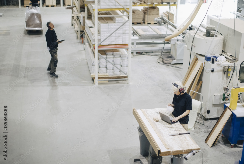 High angle view of male worker examining planks on rack while colleague using laptop at industry High angle view of male worker examining planks on rack while colleague using laptop at industry