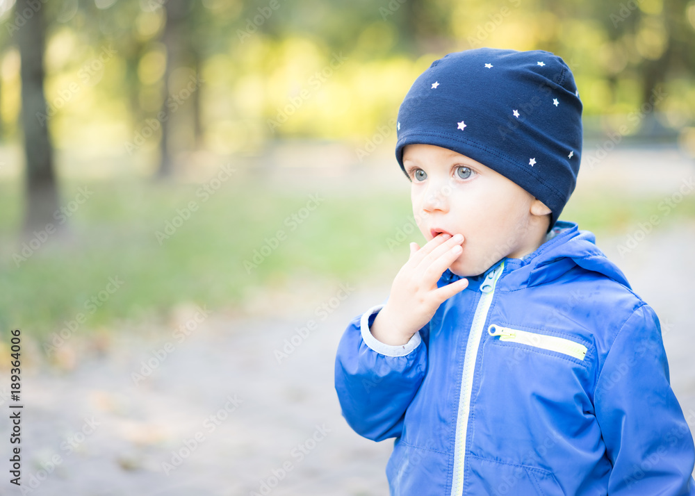Baby boy with cap at the park puts his fingers in his mouth with a perplexed face, amazed and looks into space.
