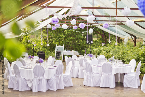Outdoor wedding in Scandinavian style in old abandoned greenhouse. White tablecloth, white chairs, green trees in background. 