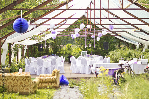 Outdoor wedding in Scandinavian style in old abandoned greenhouse. White tablecloth, white chairs, green trees in background. 