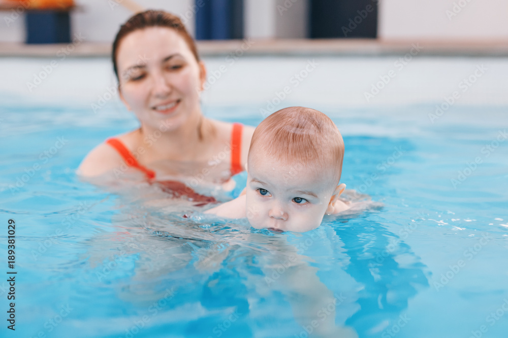 White Caucasian mother traning her newborn baby to float in swimming pool. Baby diving in water. Healthy active lifestyle. Family activity and early development concept White Caucasian mother traning her newborn baby to float in swimming pool. Baby diving in water. Healthy active lifestyle. Family activity and early development concept
