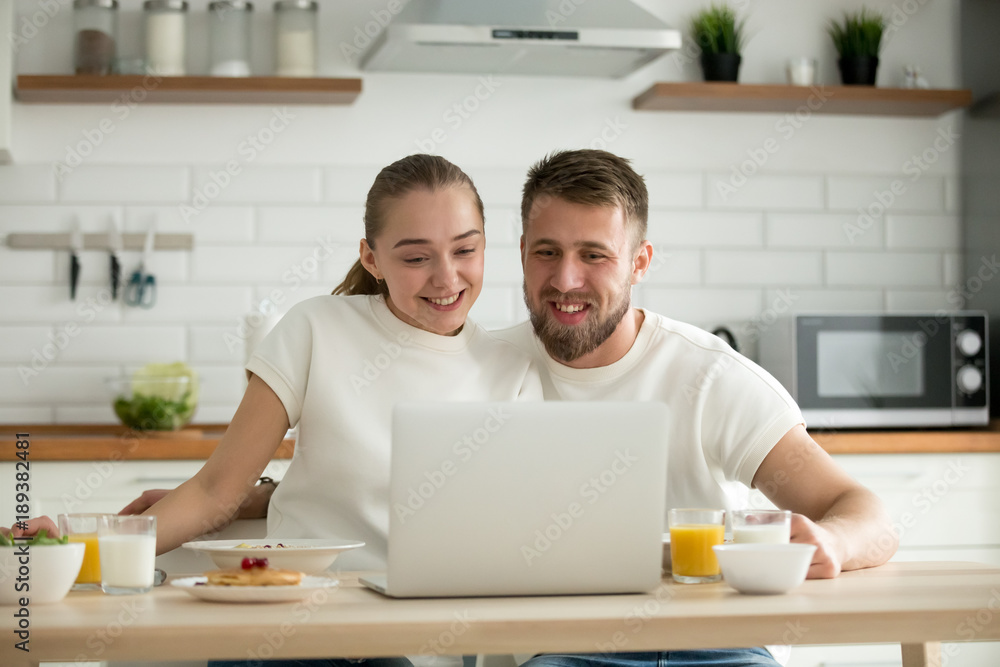Smiling happy couple enjoying breakfast in the kitchen looking at laptop screen making video call, husband and wife embracing having meal at dining table watching funny video on computer together Smiling happy couple enjoying breakfast in the kitchen looking at laptop screen making video call, husband and wife embracing having meal at dining table watching funny video on computer together