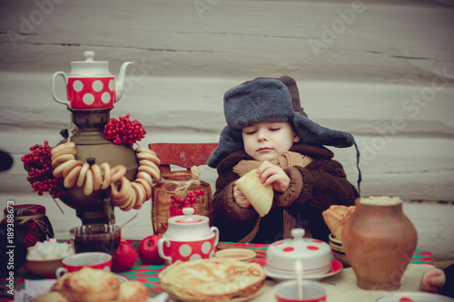 A little boy in an old fur coat and fur hat eats pancakes and jam at the holiday table. Shrovetide Maslenitsa