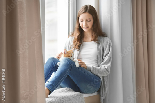 Beautiful young woman with cute cat on windowsill at home
