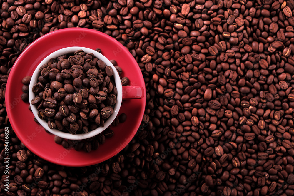 Fototapeta premium Red coffee cup and saucer one single for espresso or capuccino filled full of coffee bean on a background of scattered dark coffee beans photo top view