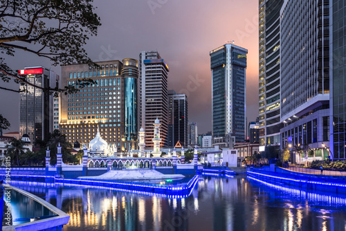 Canvas Print The office buildings reflects in the water of the Klang river in front of the Jamek mosque in the heart of Kuala Lumpur in Malaysia