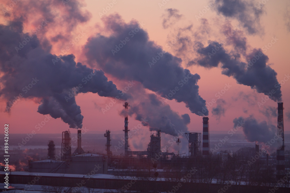 the chimneys of a refinery with smoke and steam with the pink sunset on the background