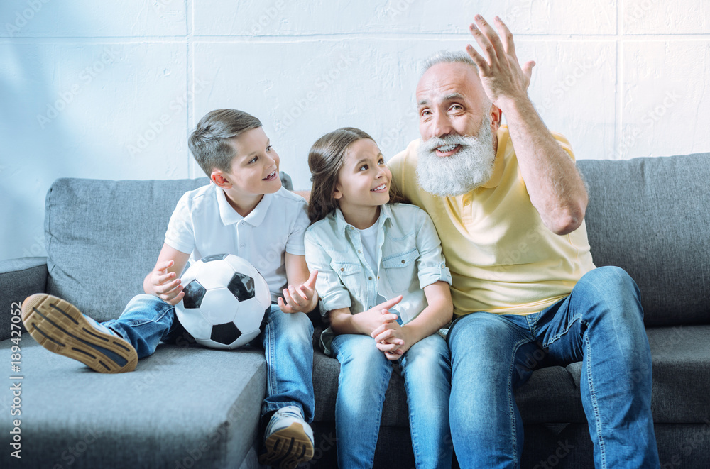 Time for a story. Adorable youngsters smiling cheerfully while sitting on a sofa and looking at their radiant granddad gesturing while telling something exciting.