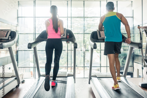 Full length rear view of a fit woman and her cardio workout, partner running on treadmills side by side during high-intensity interval training in a modern fitness club