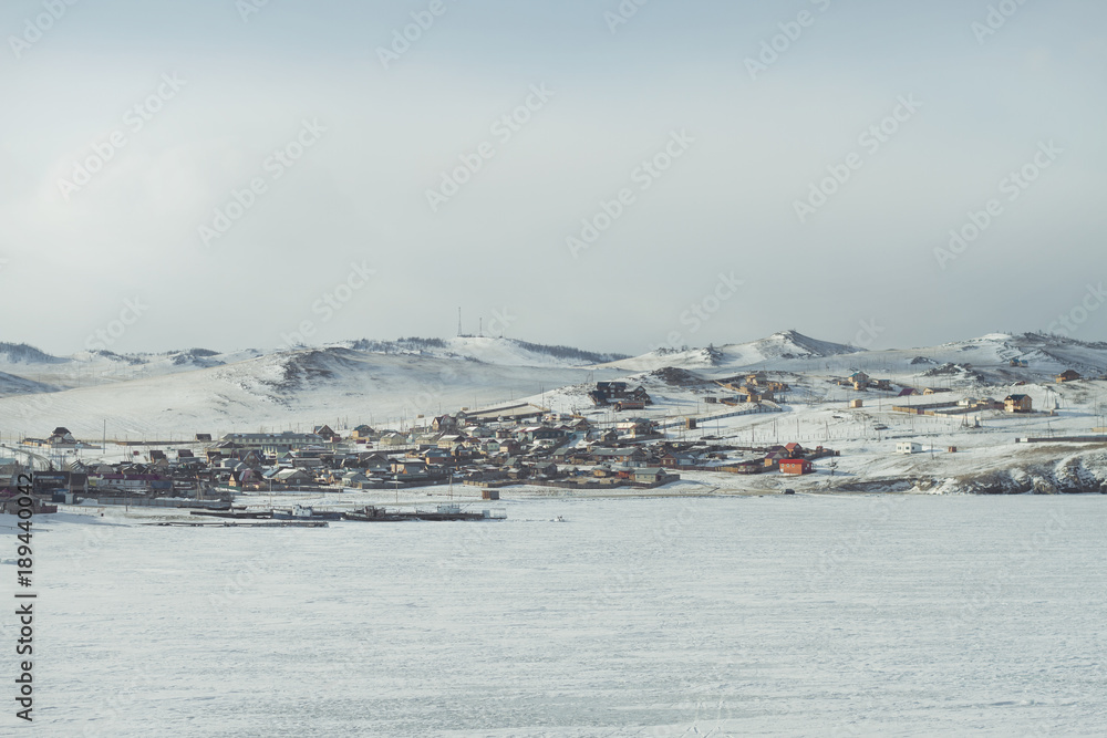 A rural landscape with houses in winter on the shore of Lake Baikal and ships on the ice in the foreground on a snowy mountainous background on a cloudy day.