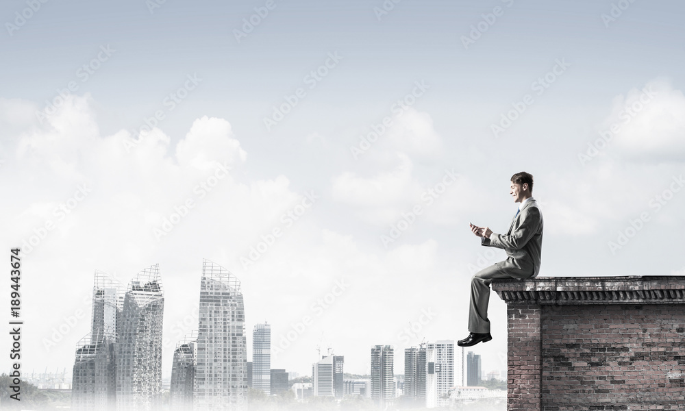 Man on roof edge send message with smartphone and cityscape at b Man on roof edge send message with smartphone and cityscape at b