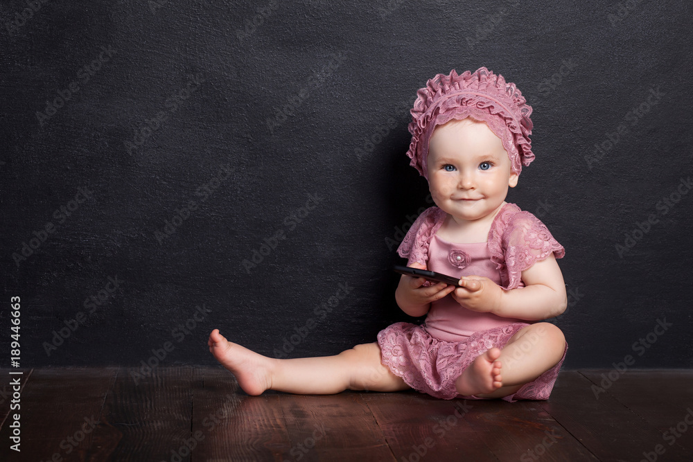 The girl sits on a brown floor and holds phone. The girl looks in the camera and smilles. Black background. The girl sits on a brown floor and holds phone. The girl looks in the camera and smilles. Black background.