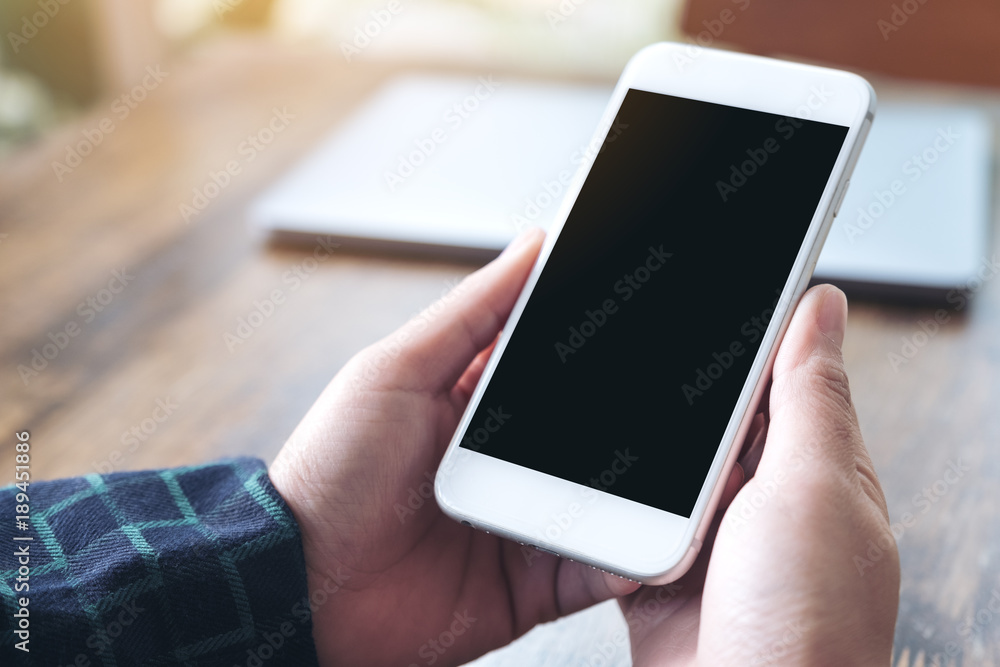 Mockup image of hands holding white smartphone with blank black desktop screen and laptop on wooden table