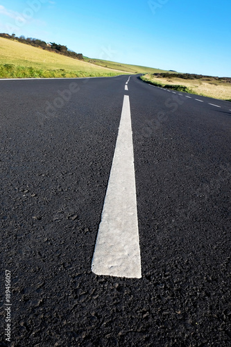 grey asphalt country road with strong white lines in the middle of the road, the road has a camber road and lines big in the foreground leading to small in the distance, green rolling hills on sides