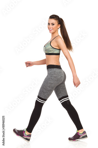 Walking Smiling Young Woman In Pink Camo Sport Bra And Striped Leggings. Side View.