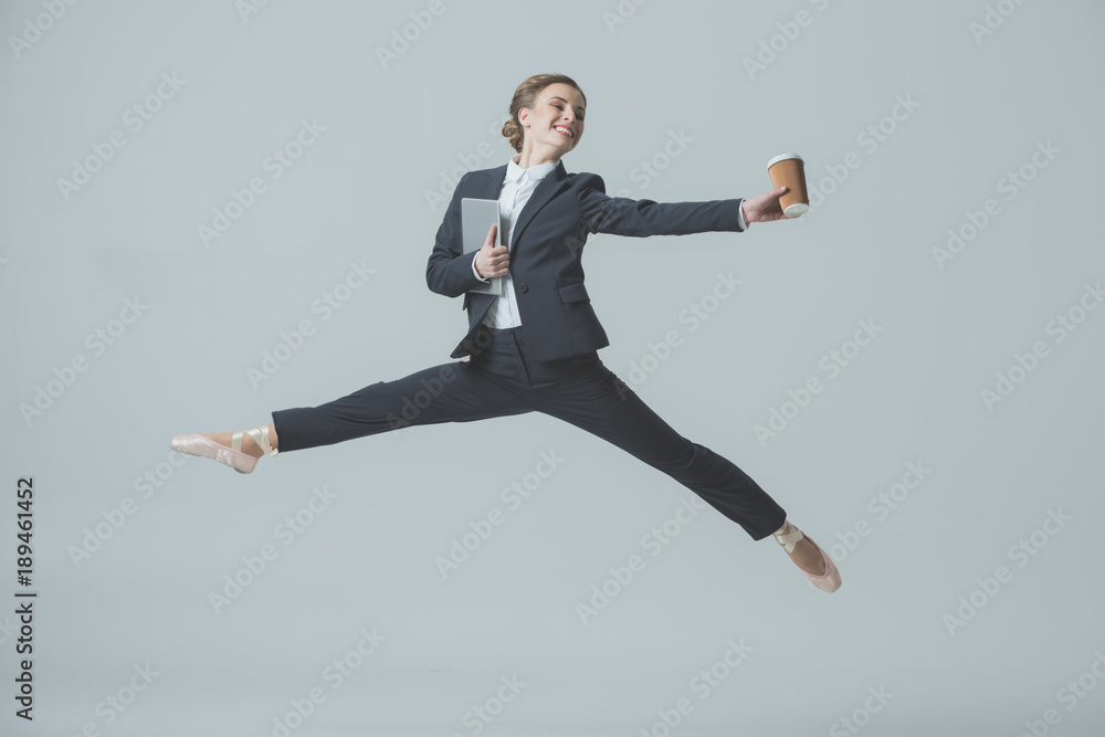 businesswoman in suit and ballet shoes jumping with coffee and tablet, isolated on grey
