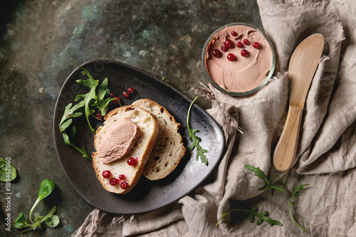 Chicken homemade liver paste or pate in glass jar with sliced whole grain bread, wood knife, cranberries, green salad served on ceramic plate with textile over dark metal background. Top view, space