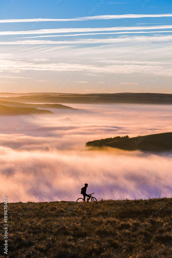 Sun setting over a fantastic Peak District mountain landscape. A biker makes its way through the hills and the fog. A calm yet exhilarating scene.