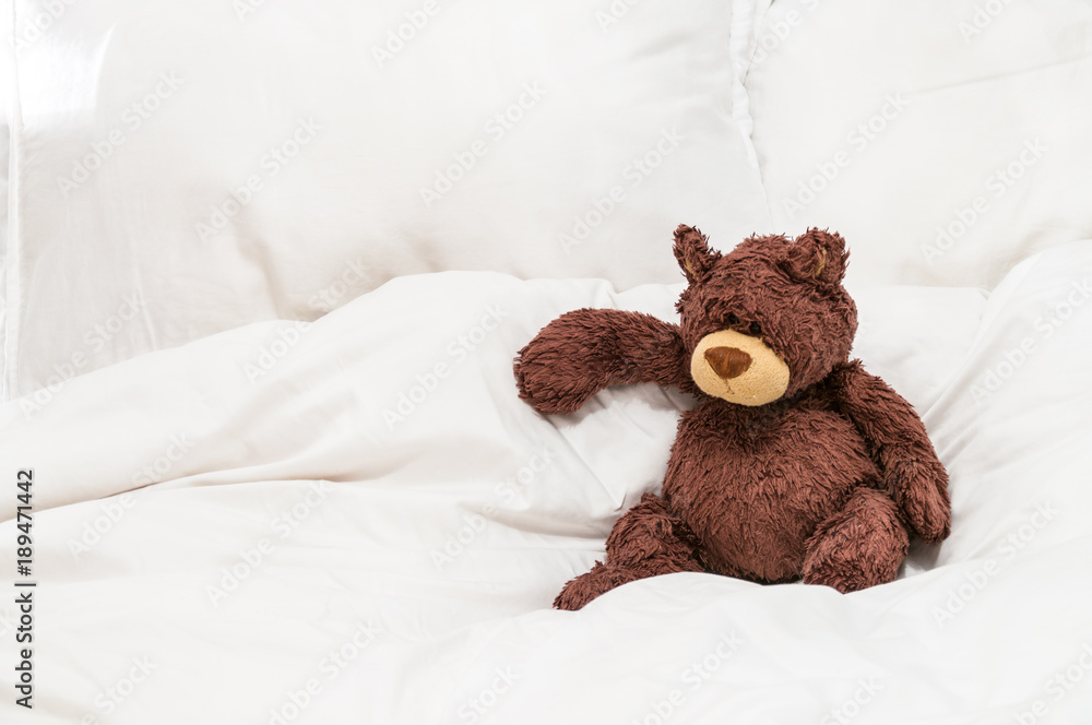 An old, worn teddy bear sat on white sheets on a bed