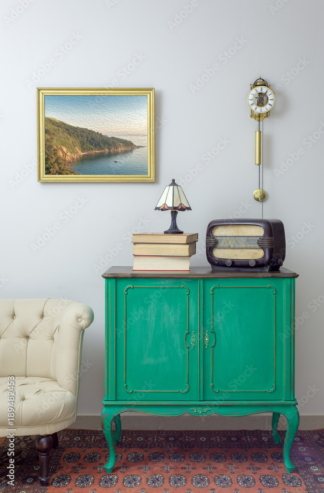 Interior shot of green vintage sideboard with old radio, stack of old  books, and table lamp on background of off white wall with hanged painting,  orange ornate carpet, and cream armchair Photos