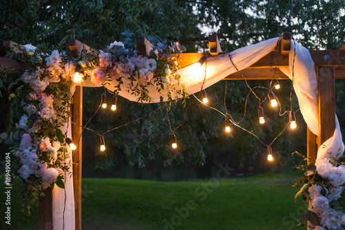 Beautiful place made with wooden square and floral decorations for outside wedding ceremony in wood. Wedding settings with electric lamp  bulbs at scenic place at night. Horizontal color photography.