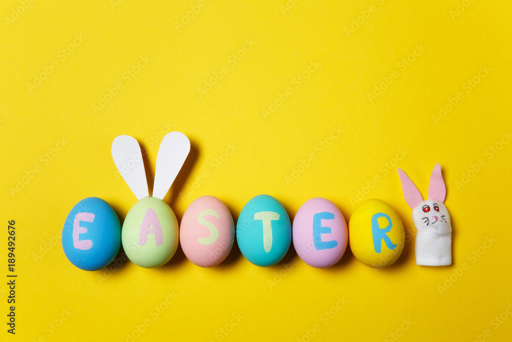 Row of colorful pastel monophonic painted Easter eggs with inscription Easter, fun bunny ears on egg, white rabbit on yellow background. Copy space for advertisement. With place for text. Top view. Row of colorful pastel monophonic painted Easter eggs with inscription Easter, fun bunny ears on egg, white rabbit on yellow background. Copy space for advertisement. With place for text. Top view.