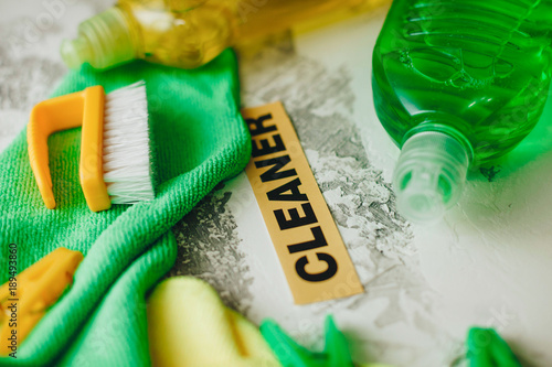 Set of yellow and green cleaning tools: rags, brushes, clothes peg and dishwashing liquid on light concrete background with cleaner inscription, top view, flat lay