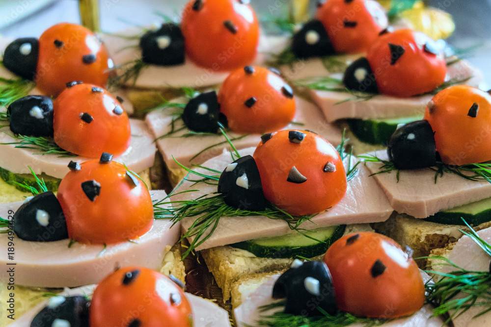 Smiling sandwiches with edible ladybugs from tomatos and olives, as well as bread, cheese, cucumber, sausage, dill. Perspective view with selective focus.