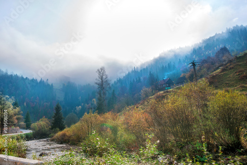 Beautiful morning on highway in mountains. Thick fog around highest mountain tops. Small river with rocks next to road. Colorful and spooky autumn background