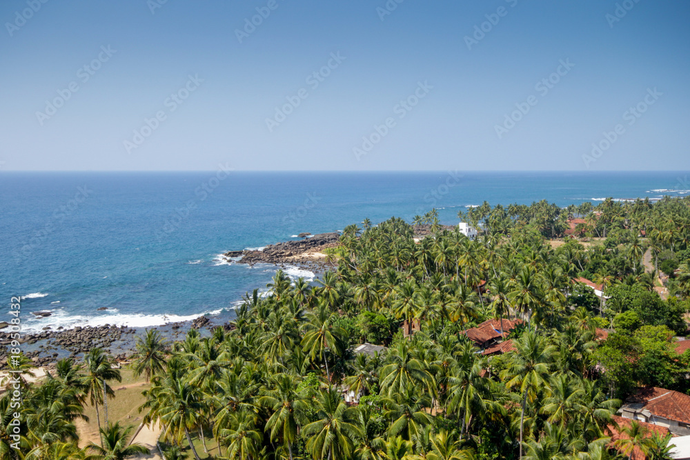 Sri Lanka. Dondra. Top view from the lighthouse to the turquoise sea (ocean), rocky beach, sandy beach, green palms, red tiled roofs of houses and boats on the jig.