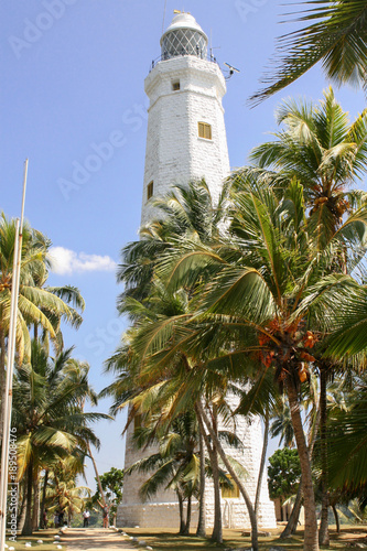 Sri Lanka. Dondra. White stone lighthouse on a stony-sandy beach among green palms. The road to the lighthouse.