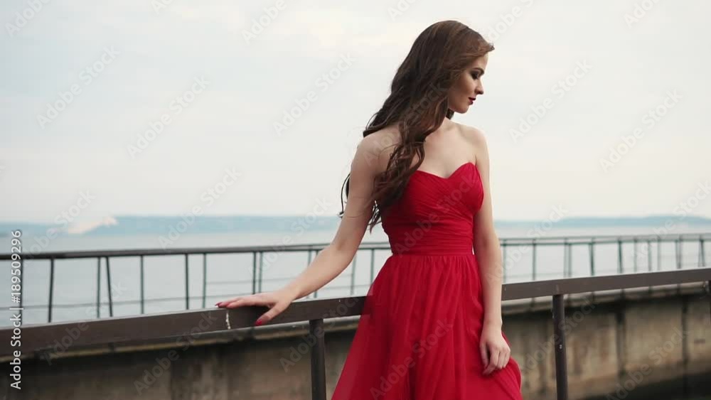 a young and pretty woman who looks gently and romantically at a pier near the ocean on a summer day, her dress fluttering under the winds, she has curly brown hair on her head a young and pretty woman who looks gently and romantically at a pier near the ocean on a summer day, her dress fluttering under the winds, she has curly brown hair on her head