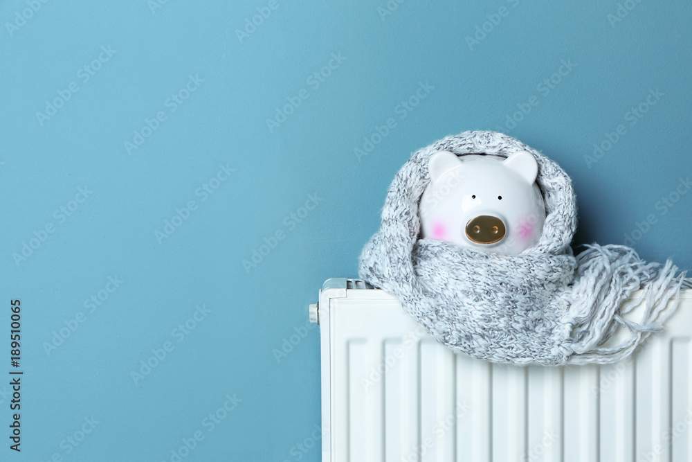Naklejka premium Piggy bank with warm scarf on heating radiator near color wall