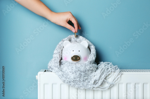 Woman putting coin into piggy bank placed on heating radiator against color background