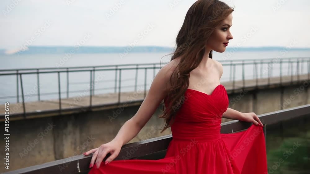 Side view shot of a gorgeous woman dressed in a red cocktail dress on the shore. She leans on the railings and looking at the horizon. Side view shot of a gorgeous woman dressed in a red cocktail dress on the shore. She leans on the railings and looking at the horizon.