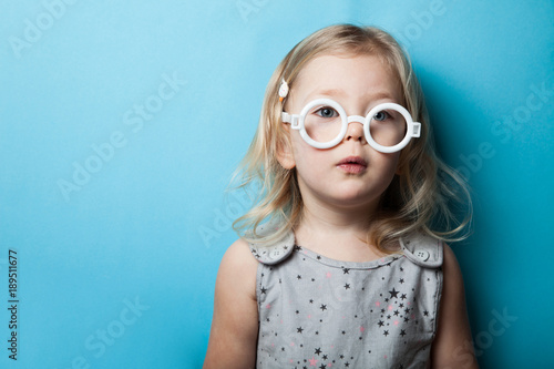 A white European little girl in toy glasses looks interested at a blue background.
