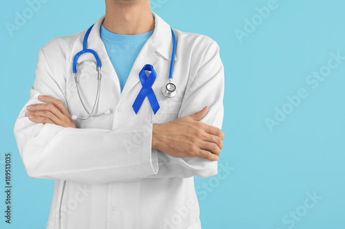 Doctor with blue ribbon on color background, closeup. Prostate cancer awareness