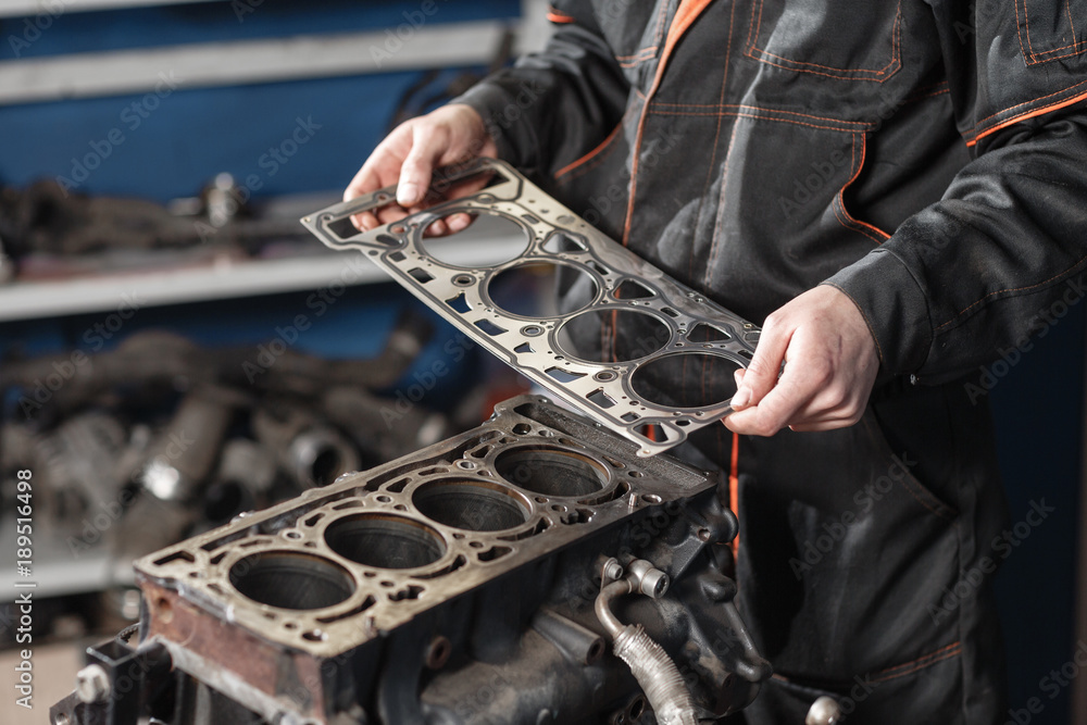 Naklejka premium Sealing gasket in hand. The mechanic disassemble block engine vehicle. Engine on a repair stand with piston and connecting rod of automotive technology. Interior of a car repair shop.